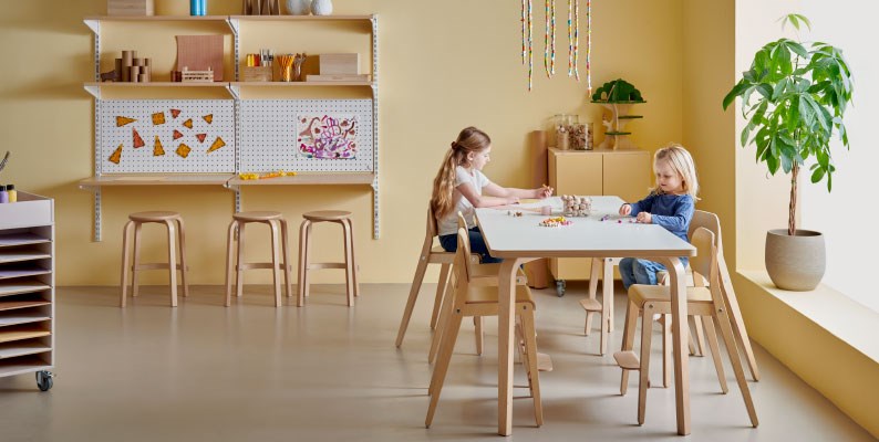 Lapset askartelevat päiväkodin ryhmähuoneessa, korkeat tuolit parantavat päiväkodin henkilökunnan ergonomiaa.